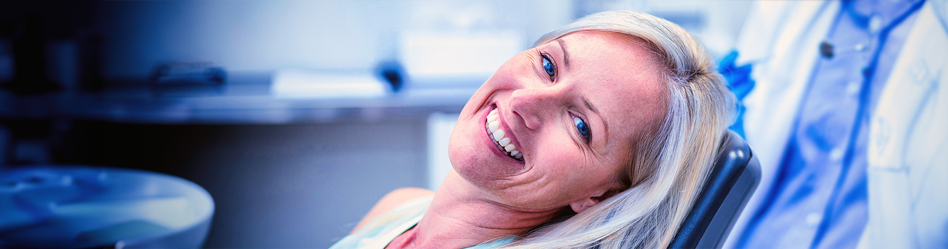 A person is seated in a dental chair, receiving care from a dental professional who stands behind them.