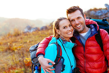 The image shows a man and woman posing together outdoors, smiling at the camera, with the man wearing a backpack and both dressed in casual hiking attire, suggesting they are on a hiking trip during the daytime.