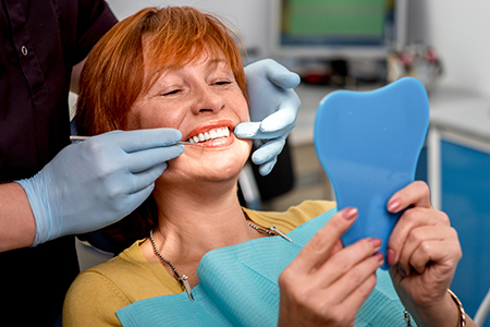 A woman wearing a surgical mask sits in a dental chair, holding a blue toothbrush, with a smile on her face.