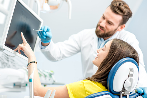 A professional setting where a dental hygienist is demonstrating a dental procedure to a patient using a computer screen, with a man standing behind them wearing a white lab coat and stethoscope.