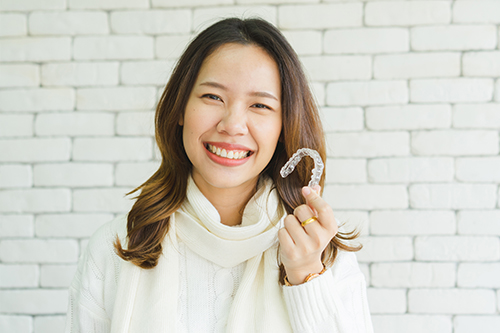 The image shows a person with a smile holding a heart-shaped object, posing against a brick wall background.