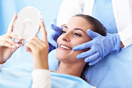 This is a photograph showing a person lying on a table with their eyes closed, receiving facial treatment from a professional who appears to be a beautician or dermatologist, using a mirror to inspect the skin.