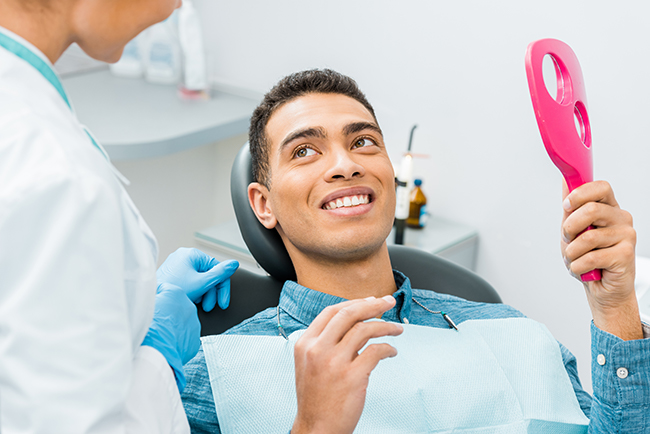 The image shows a smiling man sitting in a dental chair, holding a pink object that appears to be some kind of oral hygiene device, with a dental professional standing behind him wearing protective gloves and a mask.