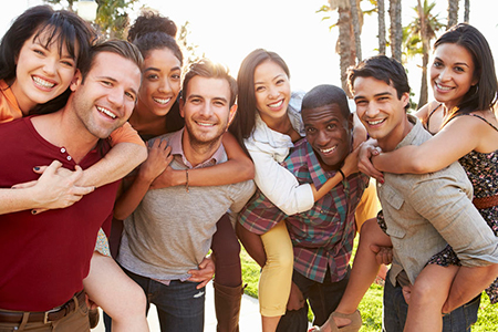 The image shows a group of young adults posing together outdoors during daylight hours, smiling and embracing each other.