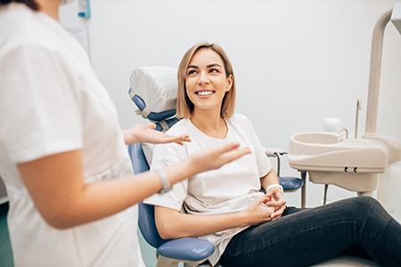 The image shows a woman sitting in a dental chair, smiling at the camera, with two dental professionals attending to her, one on each side, in a dental office setting.