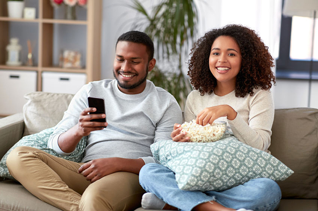 A man and woman sitting on a couch, smiling at each other while holding a smartphone and snacks, with a cozy domestic setting in the background.